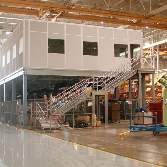 A large mezzanine structure in an industrial warehouse, featuring an enclosed office space on the upper level with windows. A wide staircase with railings leads up to the mezzanine, and the area below is used for storage and equipment, with safety fencing surrounding the ground floor.