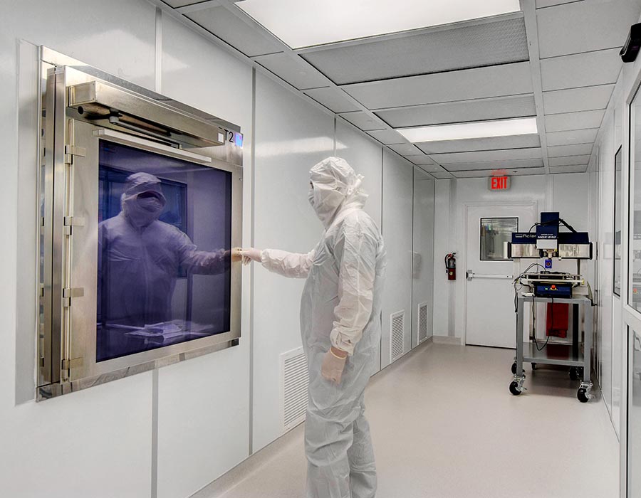 Technician in full cleanroom attire working inside an Allied Modular cleanroom with integrated pass-through chamber and wall-mounted equipment.