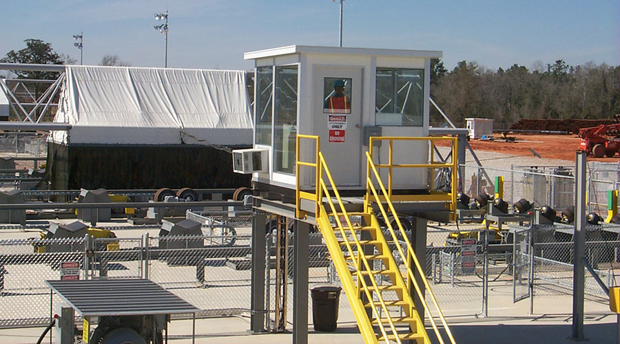 A modular observation tower with a white booth and yellow stairs in an industrial site, with a worker inside the booth and various industrial equipment visible in the background.