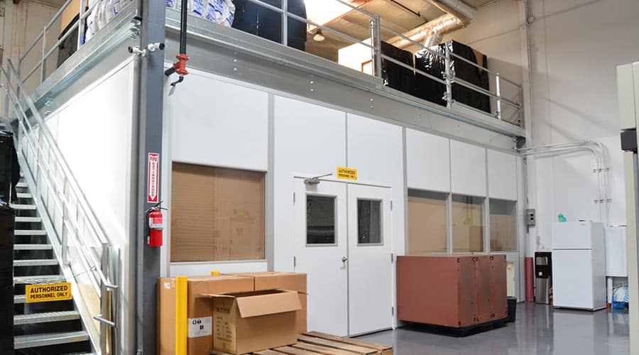 A modular office structure inside a warehouse, with an overhead mezzanine and stairs for additional storage or workspace.