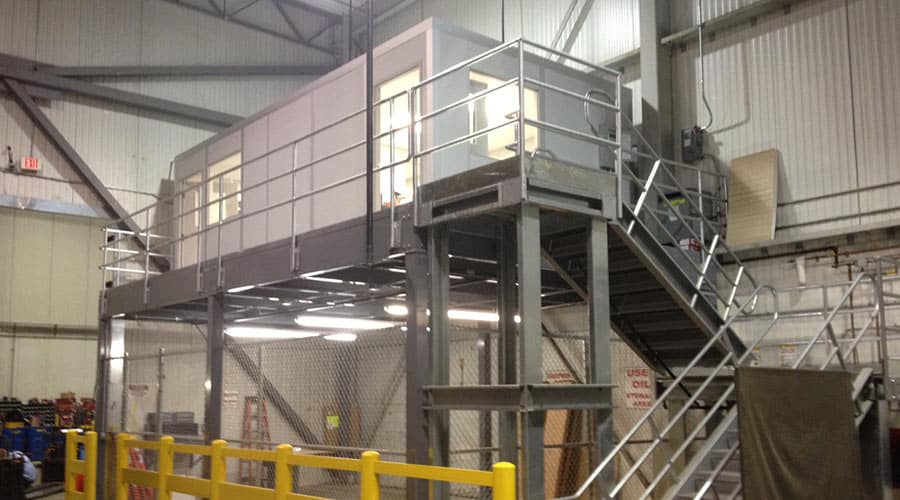 A modular mezzanine office space elevated above a workshop area with metal stairs and railings.