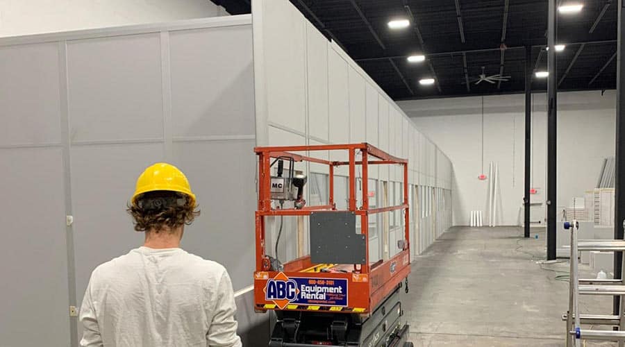 A man in a yellow hard hat stands next to orange scissor lift next to a modular office enclosure.