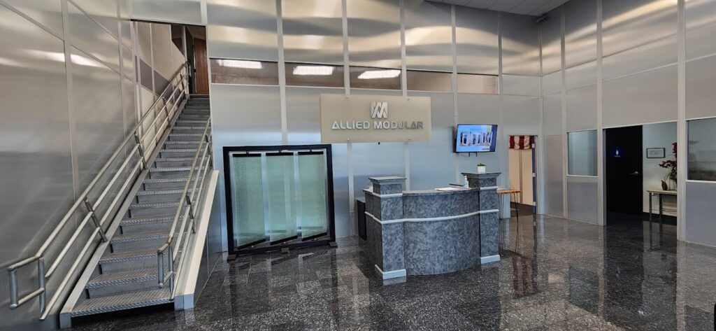 Modern office lobby with Allied Modular branding, featuring modular walls, a reception desk, and a staircase with metal railings.