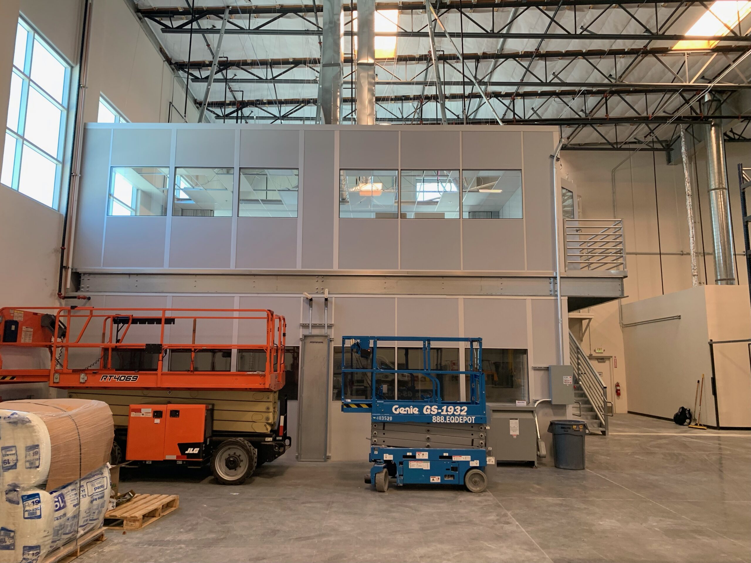 Two-story modular factory office with large windows and a staircase, situated inside an industrial warehouse, alongside scissor lifts for easy access and mobility.