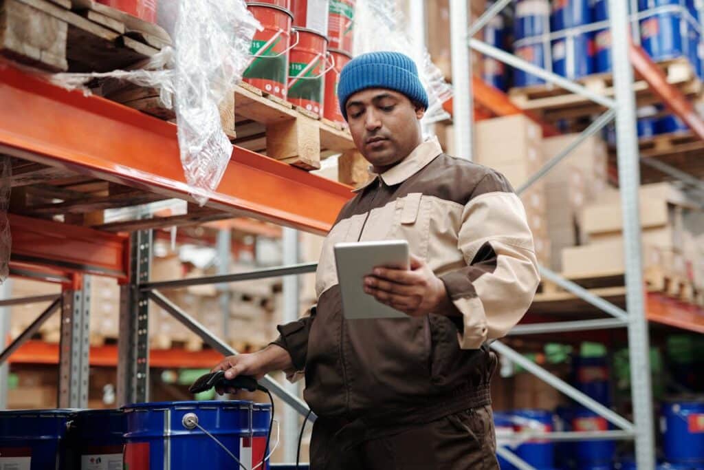 A warehouse worker using a tablet to manage inventory while standing next to neatly stacked shelves.