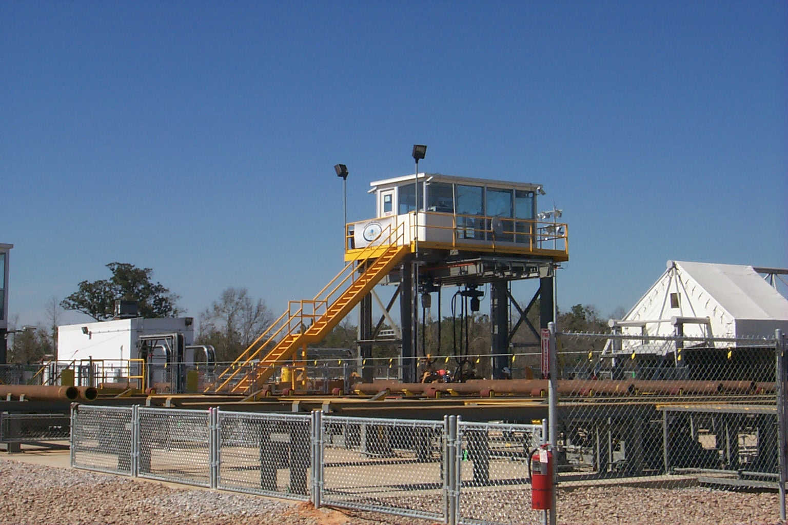 A tall modular observation tower in an industrial setting, featuring a white booth with large windows, supported by a steel structure with yellow stairs and railings.