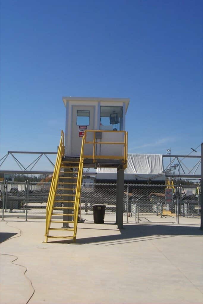 A modular observation tower with a white booth and yellow stairs, situated in an industrial area under a clear blue sky.