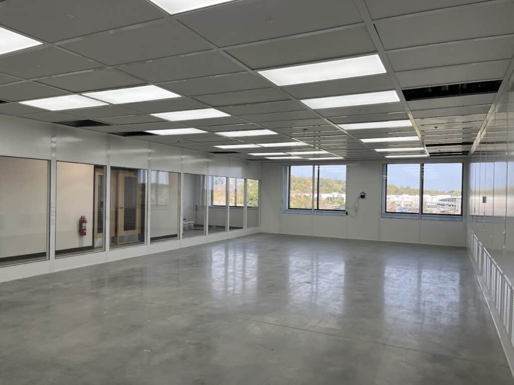 Spacious modular office interior with large windows, white panel walls, and a polished concrete floor, featuring overhead lighting and views of the outside.
