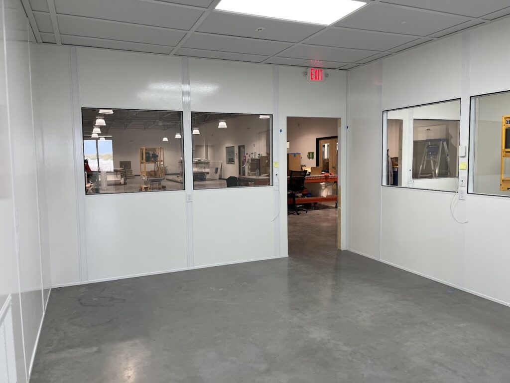 Interior view of a modular warehouse office with white panel walls, observation windows, and a door leading to the main warehouse.