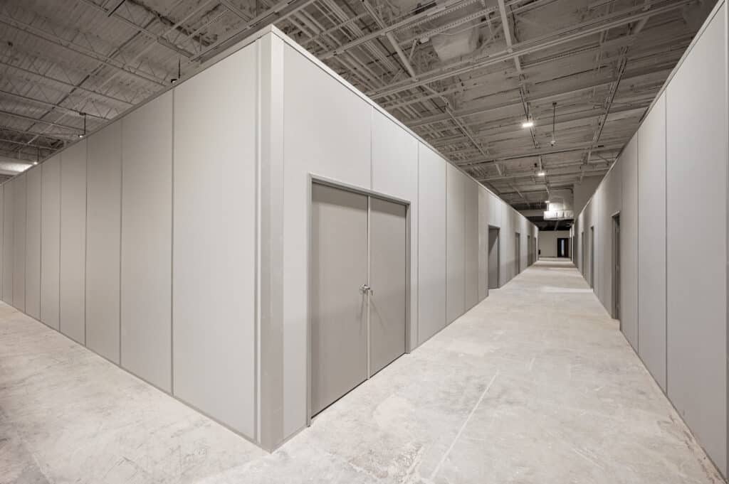 A warehouse interior featuring temporary modular walls forming separate rooms with closed doors. The space has an industrial look with exposed ceiling beams, lighting fixtures, and an unfinished concrete floor. The modular partitions provide a clean and organized layout for adaptable workspace solutions.