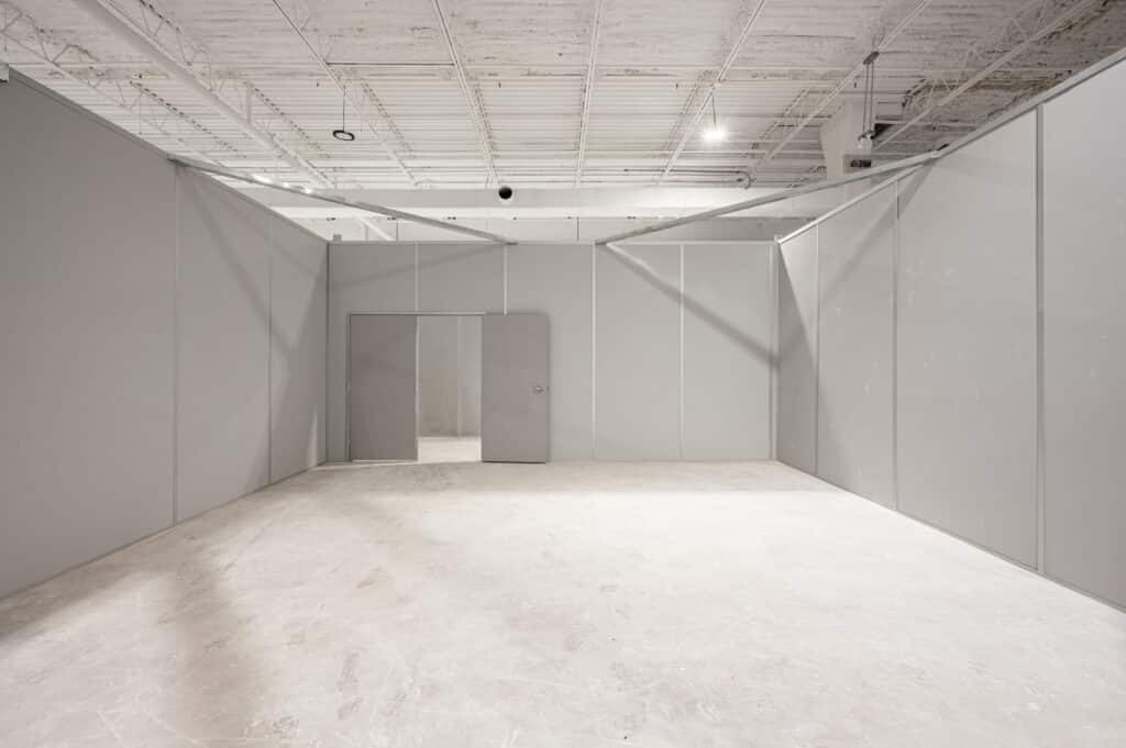 A warehouse space featuring temporary modular walls in a neutral gray color, with an open doorway leading to another section. The ceiling includes exposed beams, lighting fixtures, and ductwork, while the concrete floor appears unfinished.