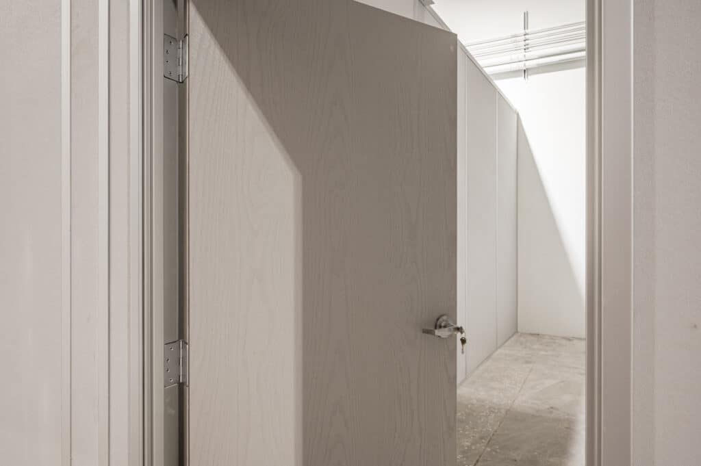 A close-up view of a partially open door leading into a room constructed with temporary modular walls. The space features a neutral color scheme, with exposed ceiling pipes and an unfinished concrete floor, highlighting the practicality and adaptability of modular partition systems.