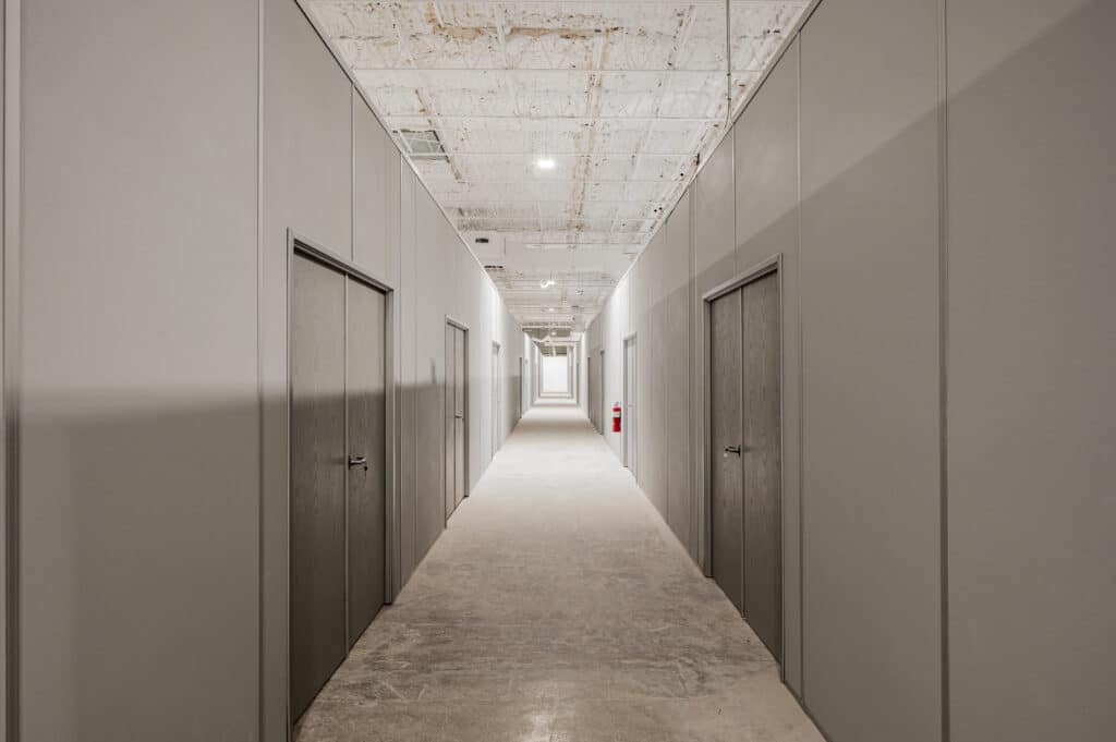 A long hallway in a warehouse featuring modular wall dividers with doors, creating separate rooms for storage or office use.